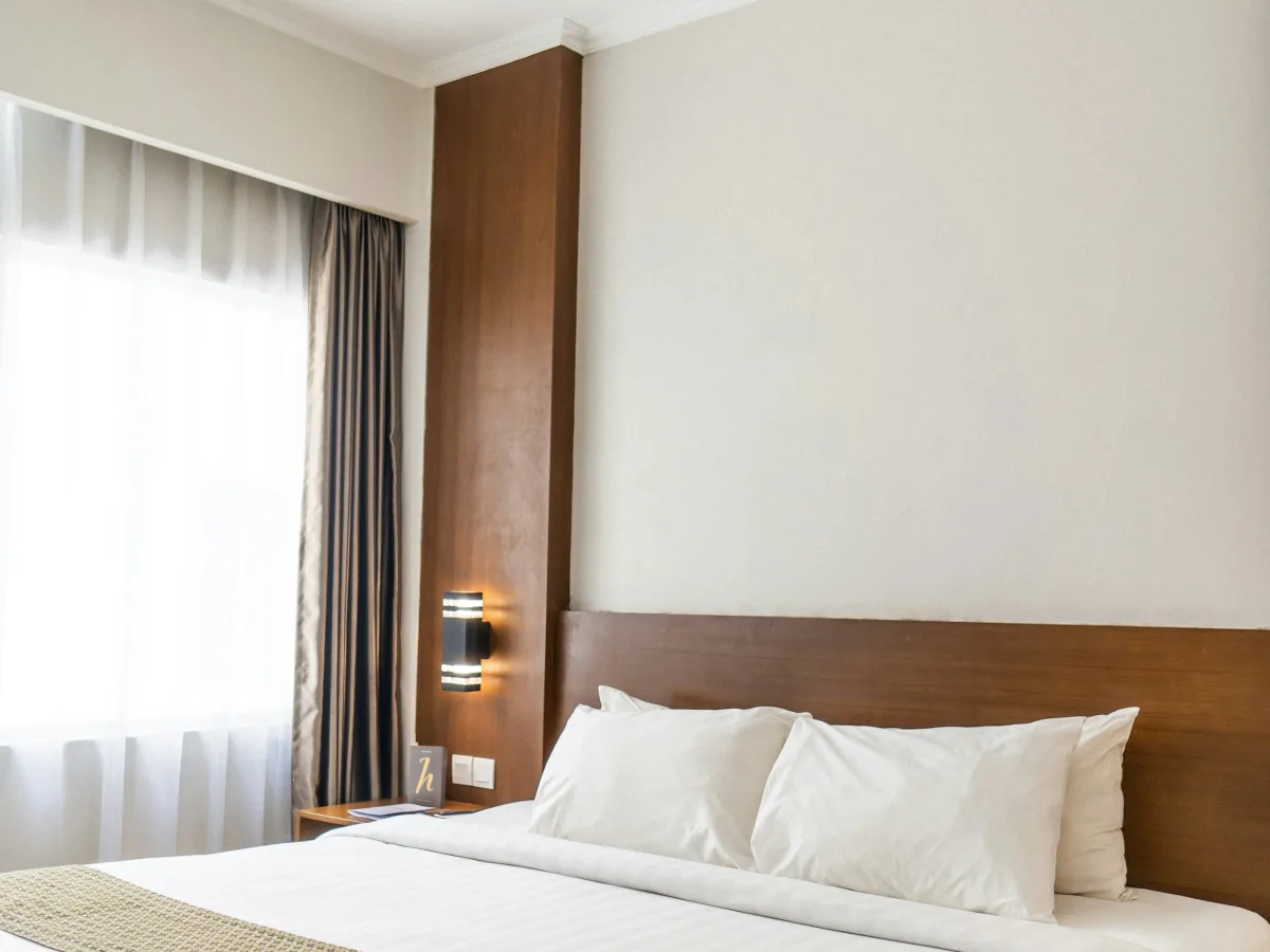 Giường đôi ga trắng, đầu giường gỗ sáng màu, cửa sổ kính có rèm xám